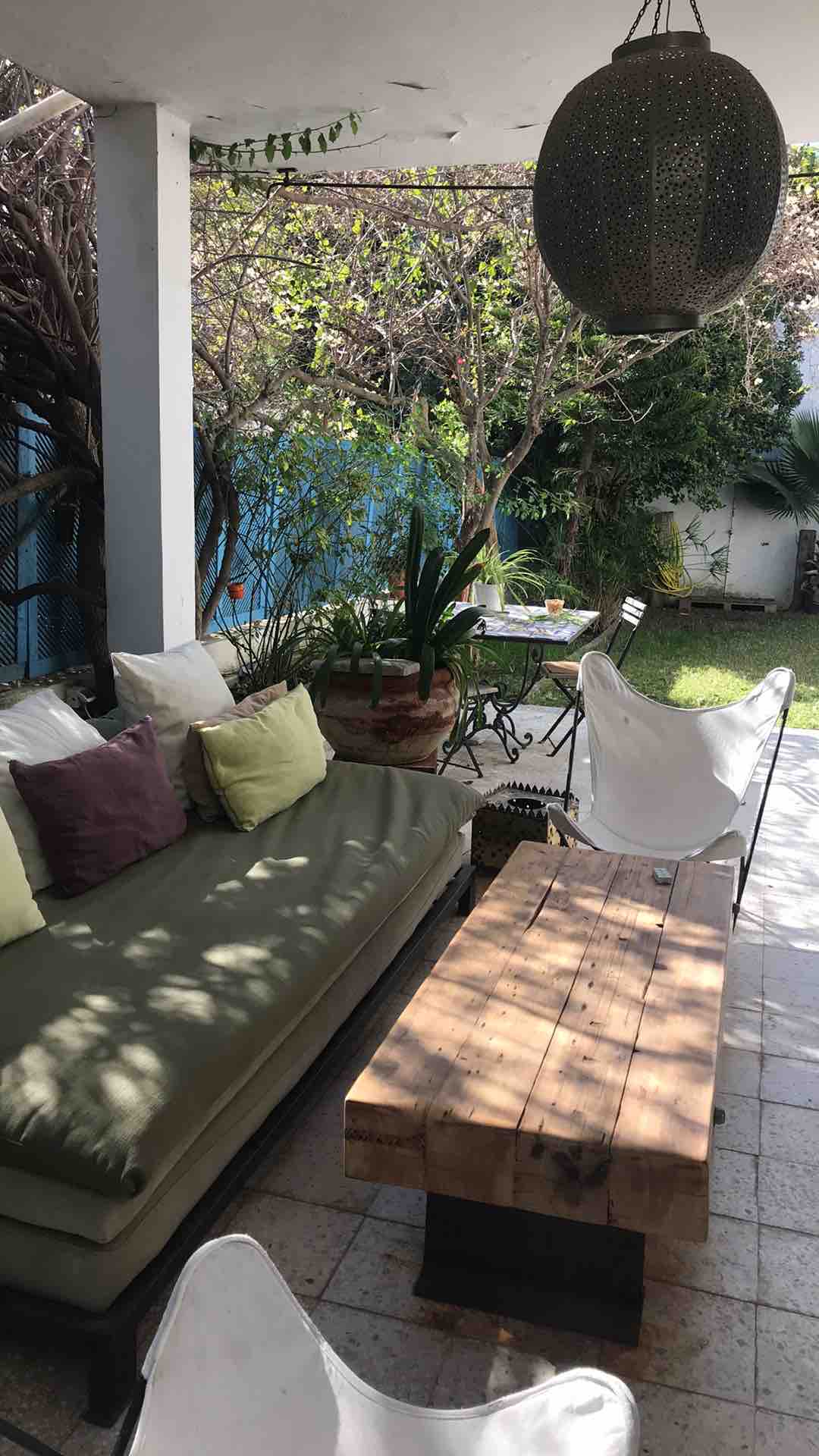 A shaded terrace is depicted with a comfortable, cushioned seating area featuring a vibrant mix of decorative pillows. A rustic wooden table rests in front, complemented by contemporary chairs. Lush greenery surrounds the space, enhancing the inviting atmosphere of the outdoor area.