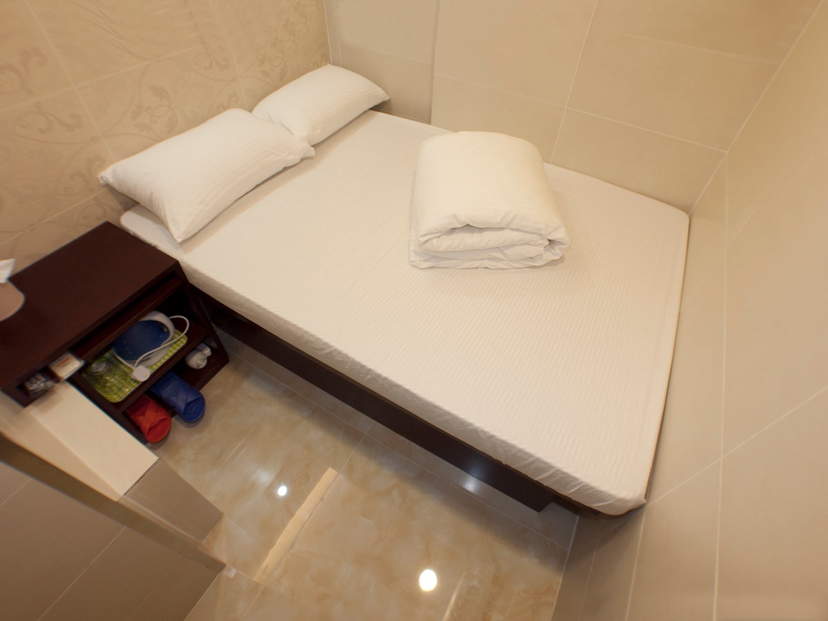 A compact sleeping area is seen with a spacious bed dressed in crisp white linens and two neatly arranged pillows. A small nightstand is located next to the bed, holding a few items, while polished tiles reflect light in the room.
