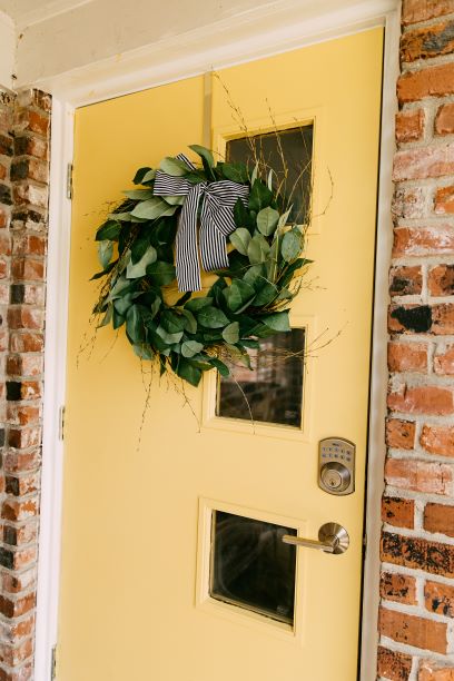 A bright yellow door is adorned with a circular wreath made of greenery and accented with a black and white striped ribbon. Two small windows, framed by brick, complement the welcoming entrance.
