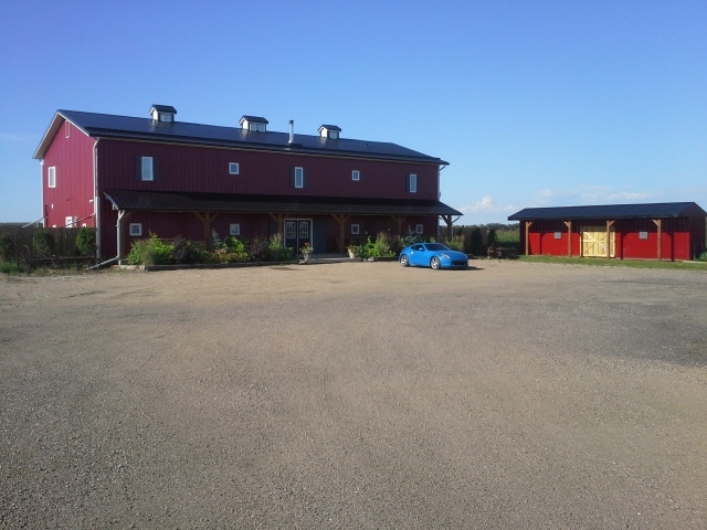 A large, red barn-style building with a metal roof is set against a clear blue sky. A gravel parking area is visible in the foreground, with a blue car parked nearby. A smaller red structure is situated to the right of the main building.