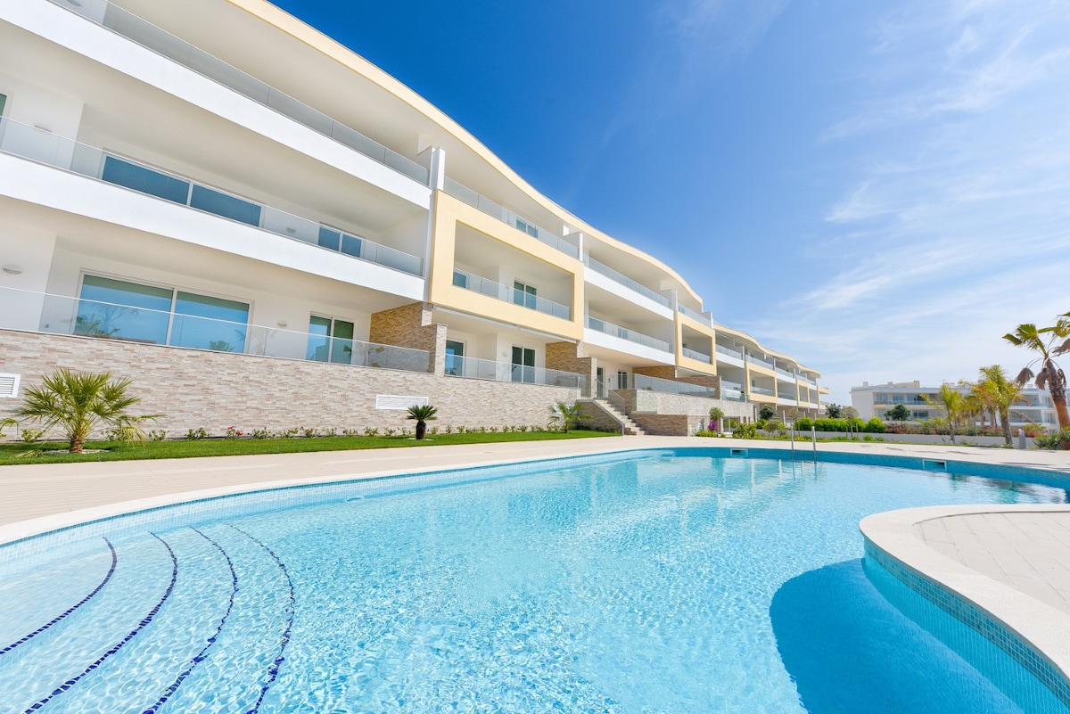 The image shows a modern condominium building with multiple balconies overlooking a freeform pool. The pool area features a curved edge and is surrounded by landscaped gardens and palm trees. A clear blue sky adds to the inviting atmosphere of the outdoor space.
