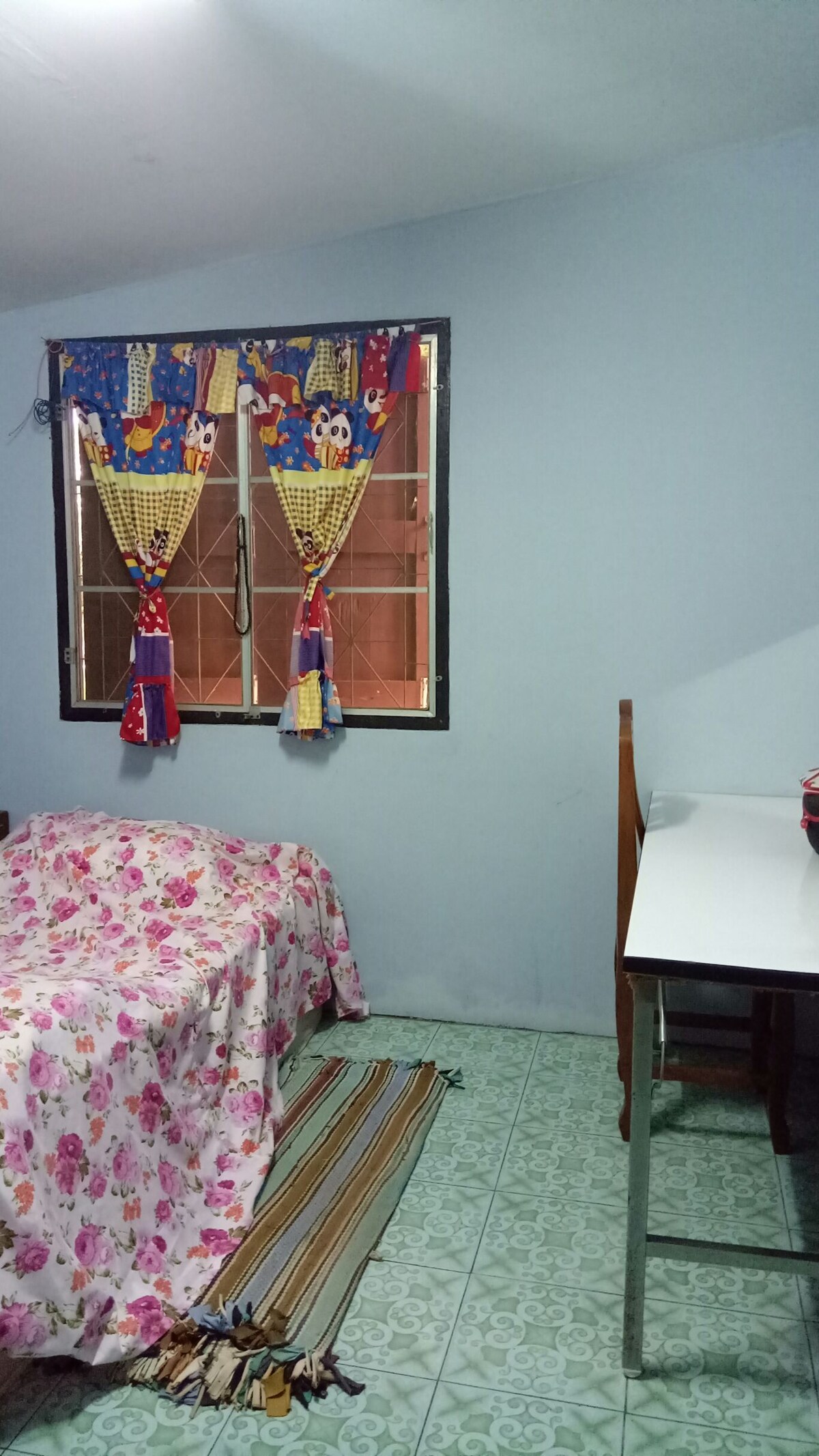 A modest sleeping area is featured, with a floral-patterned bedspread covering a spacious bed. A small table sits beside it, accompanied by a colorful curtain over a window that allows natural light to enter. The tiled floor adds a touch of color to the room.