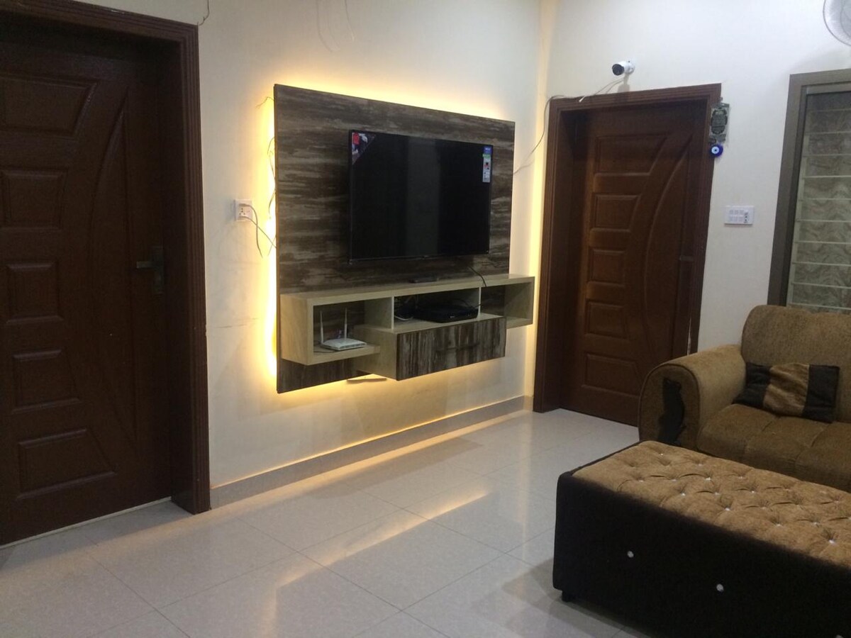 The living room features a modern entertainment unit with a flat-screen television mounted on a wooden wall panel. A comfortable sofa with a neutral tone is positioned nearby, while warm accent lighting highlights the space. Two wooden doors lead to different areas of the home.