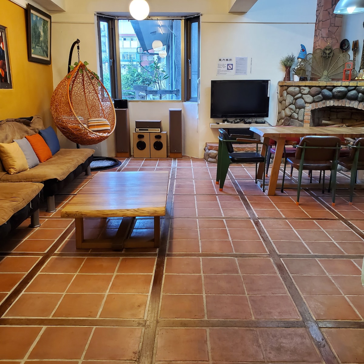 A common area is shown with a textured tile floor. A cozy sofa with colorful pillows is positioned near a large window, alongside a hanging rattan chair. A wooden coffee table and a dining table with chairs are visible, and a television is located next to a stone fireplace.