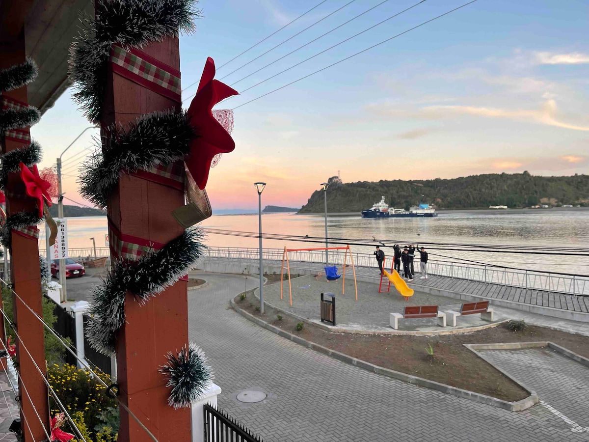 A view from a porch shows a waterfront scene, featuring a playground with swings and a slide. In the background, a hillside is visible alongside the bay, with boats anchored in the water. Decorative holiday garlands are seen adorning the railing.