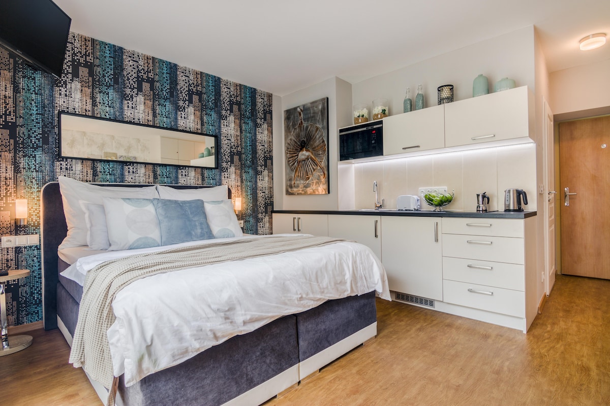 A modern sleeping area features a spacious bed layered with soft linens and decorative pillows. A kitchenette with sleek white cabinetry is visible, equipped with essential appliances. Stylish wall art and a large mirror enhance the room's character, while warm wood flooring adds to the overall ambiance.