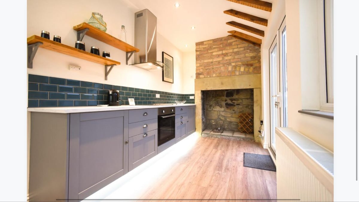A modern kitchen showcases sleek cabinetry and a contrasting tiled backsplash. Natural light streams in through the windows, illuminating the spacious counter area. A feature fireplace made of stone is visible, adding character to the room. Open shelving displays neatly arranged items.
