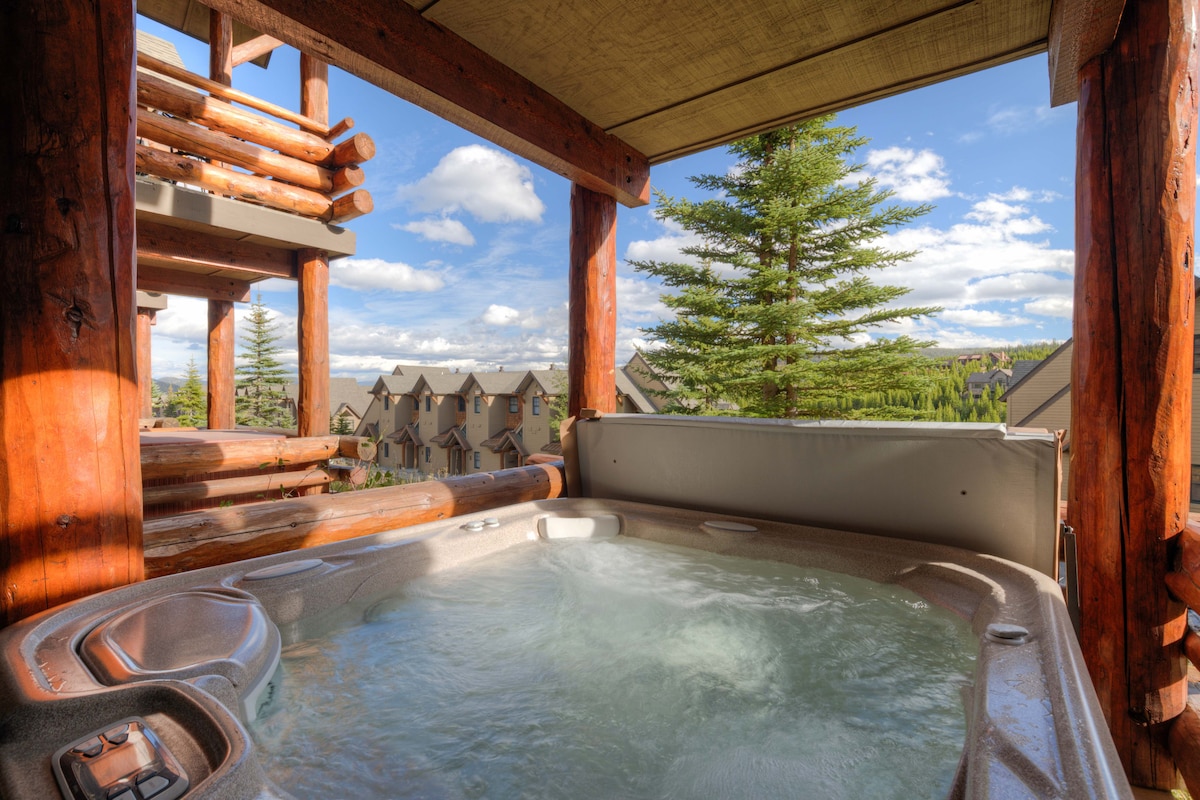 A private hot tub situated on a wooden deck offers views of the surrounding mountains and trees. The spa features a smooth, curved design with bubbling water, surrounded by sturdy log pillars that frame the picturesque outdoor setting.