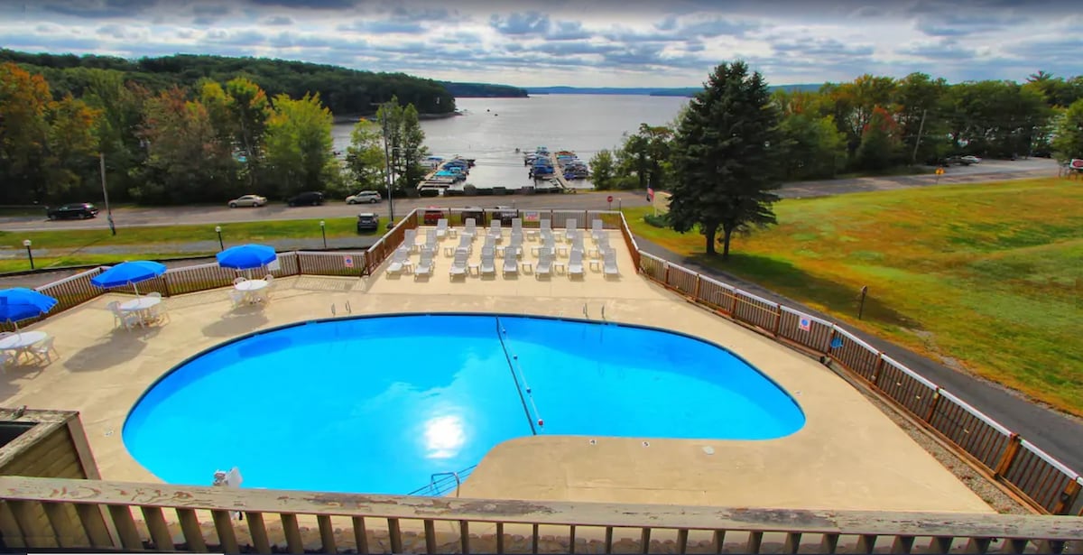 An outdoor pool is surrounded by lounge chairs and umbrellas, offering a space for relaxation. Beyond the pool area, views of Lake Wallenpaupack are visible, framed by greenery and boats moored along the shoreline.