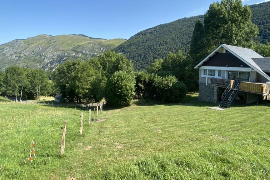 A chalet is situated amidst lush greenery, framed by rolling hills and mountains in the background. The exterior features a blend of stone and wood, with a spacious deck and large windows allowing for ample natural light. The surrounding landscape offers a serene, open atmosphere.