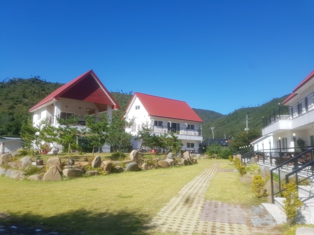 Two multi-story buildings are positioned against a clear blue sky, showcasing red-tiled roofs. A well-manicured lawn with smooth stone pathways and scattered greenery connects the structures, adding to the serene setting framed by rolling hills.