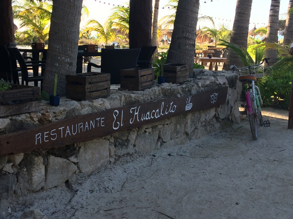 The outdoor area of a restaurant is shown, featuring a stone wall with the name 'El Huacalito' painted in bold letters. Tables and chairs are arranged among palm trees, and a bicycle is propped against the wall, contributing to a relaxed beach atmosphere.