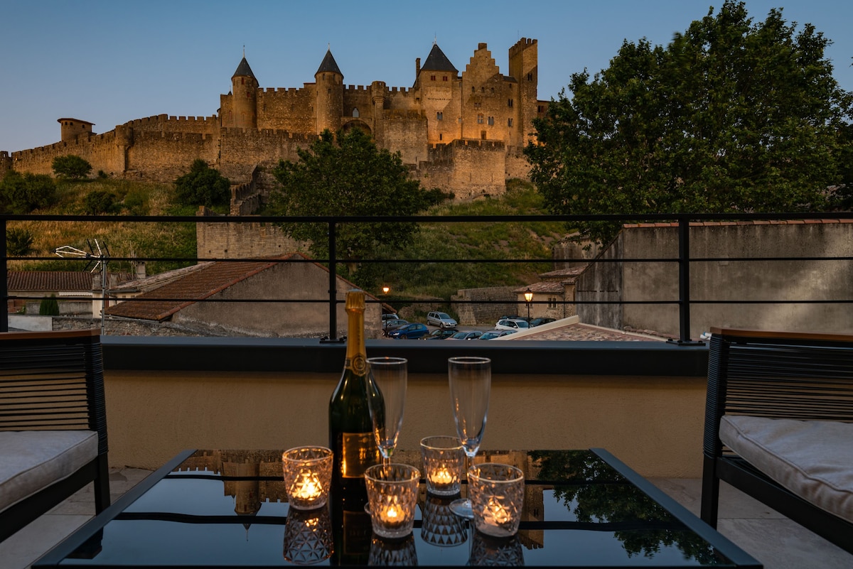 A terrace is seen, featuring a glass table set with champagne flutes and lit candles. In the background, the medieval fortress stands majestically against a twilight sky, highlighting the historic architecture and surrounding greenery.
