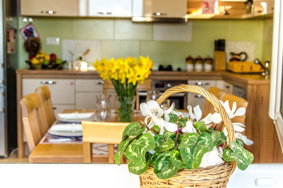 A cozy kitchen area features light wooden cabinetry and a dining space set for four. A basket of white flowers and greenery is positioned in the foreground, while yellow daffodils are seen on the counter. Soft lighting enhances the welcoming atmosphere.