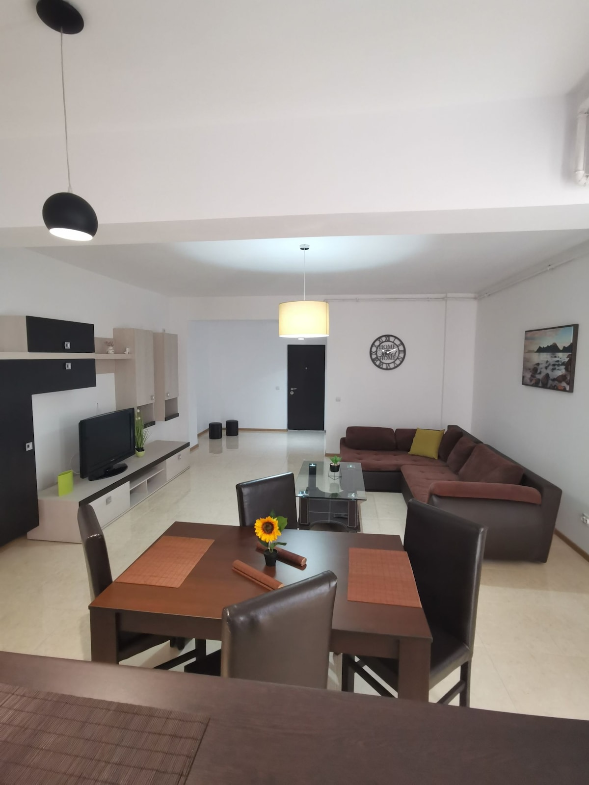 An inviting living area features a brown sectional sofa, a sleek television unit, and a modern coffee table adorned with a sunflower. A dining table with place settings is visible, while ambient lighting is provided by a pendant lamp above. Neutral wall colors create a spacious feel.