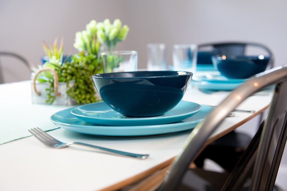 A neatly arranged dining table features blue and turquoise dishware. A large blue bowl is centered on a matching plate, complemented by silver utensils. Clear glasses and a small, green plant centerpiece enhance the simple, modern aesthetic.