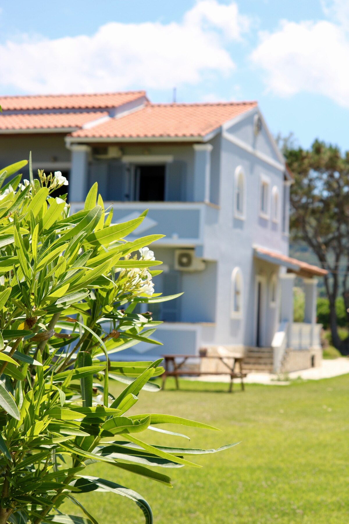 A seafront apartment is framed by lush green foliage. The building, painted in soft blue, features arched windows and a tiled roof. A well-maintained garden area lies in front, complemented by clear blue skies and scattered clouds.