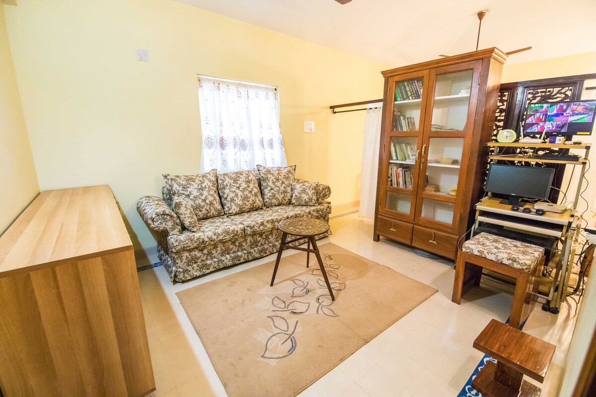 A cozy sitting area is presented with a floral-patterned sofa and a small round table. A wooden cabinet displays books and decorative items. A television setup is visible, complemented by a light-colored rug underfoot and a window draped with sheer curtains, allowing natural light to filter in.