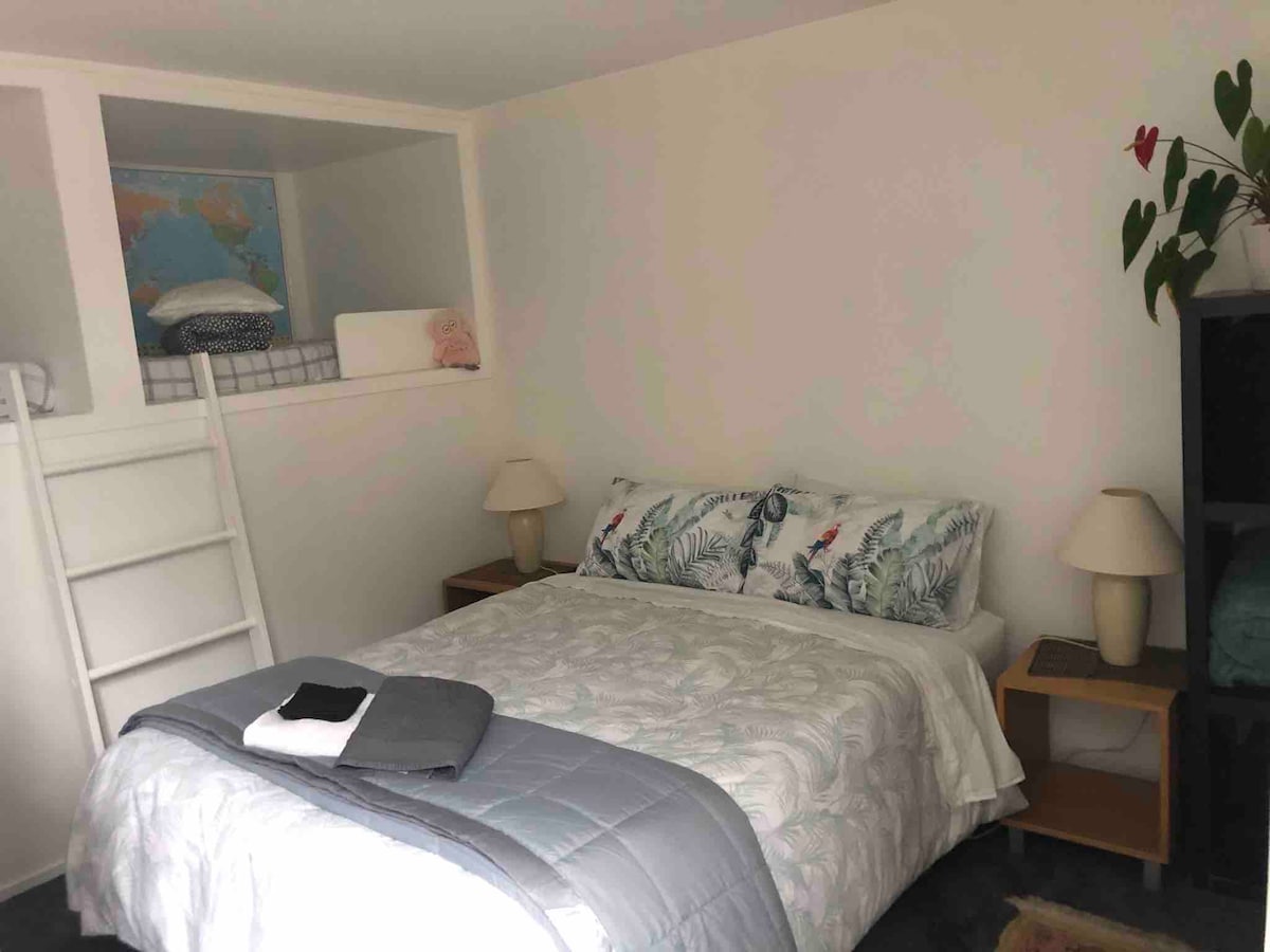 A cozy bedroom is depicted, featuring a queen bed with light-colored bedding and decorative pillows. A small bedside table is located on each side of the bed, with lamps providing soft light. Above the bed, two cubby beds are accessible, suitable for children.