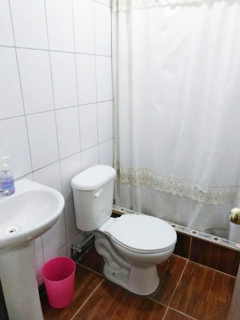 The bathroom features white tiled walls with a shower curtain adorned with lace. A pedestal sink is positioned next to a modern toilet, and a pink waste bin is included on the floor, offering convenience and functionality in the space.