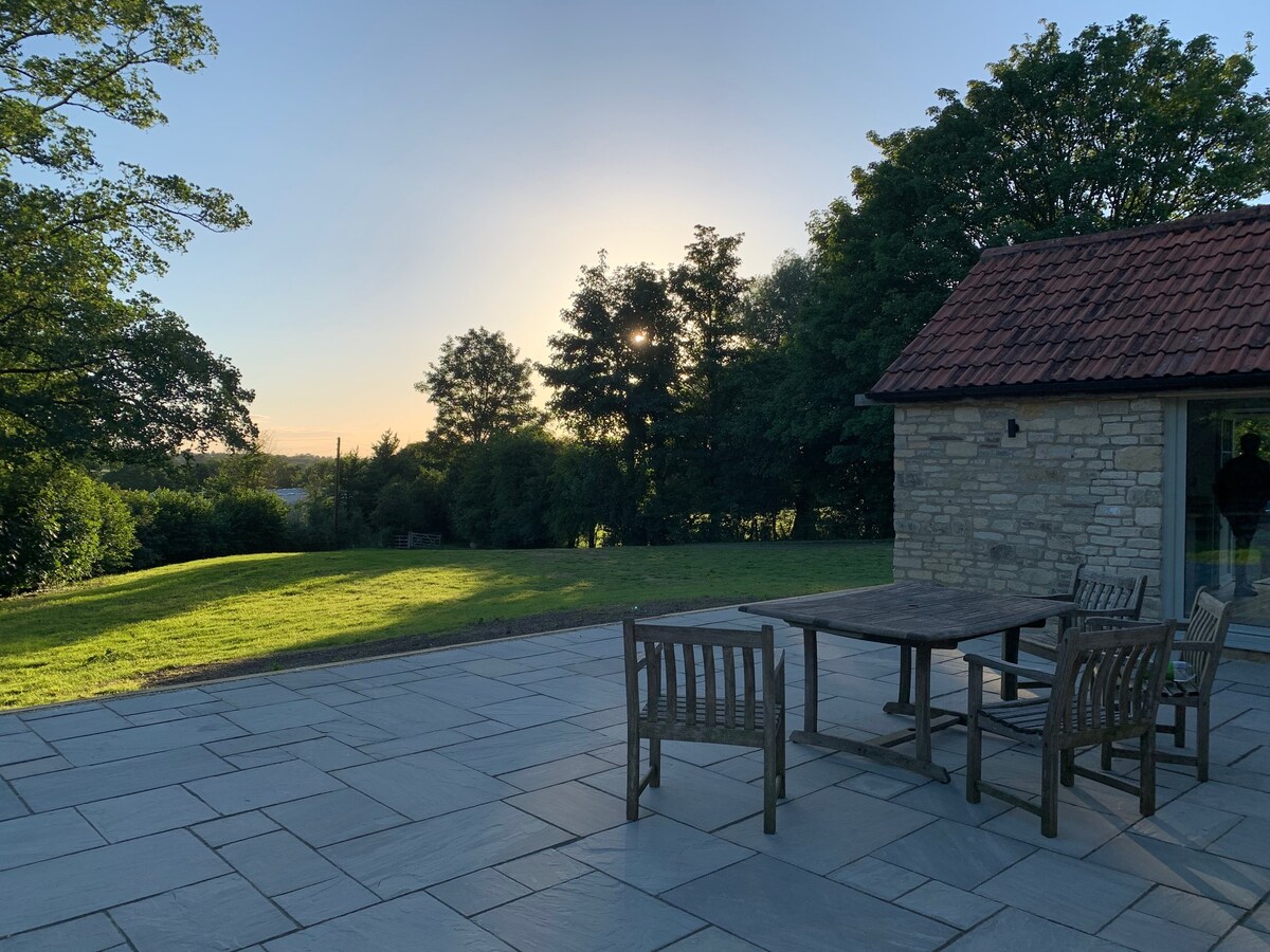 A stone patio area features a wooden table surrounded by four chairs, set against a backdrop of greenery and trees. The sun is seen setting on the horizon, casting a warm light across the open space.