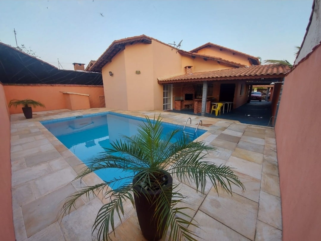 A well-maintained outdoor area features a rectangular swimming pool surrounded by light-colored stone tiles. A tall plant in a dark pot adds greenery. The house, with a terracotta exterior and tiled roof, has an open layout visible through an adjacent space.