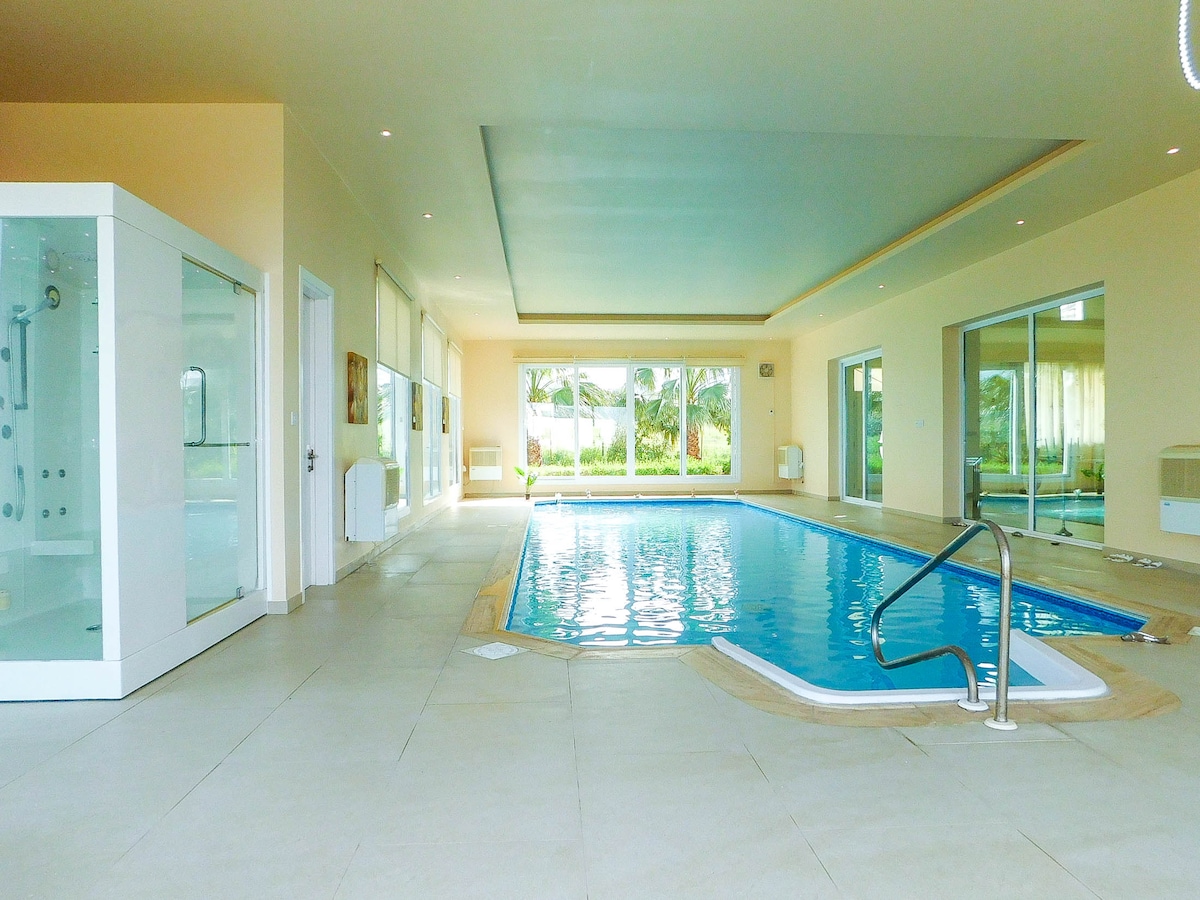 An indoor heated pool is featured within a spacious area, surrounded by large glass windows that provide a view of the outdoor greenery. Light-colored walls and tiled floors create a bright and airy atmosphere. In the corner, a modern shower facility is visible.