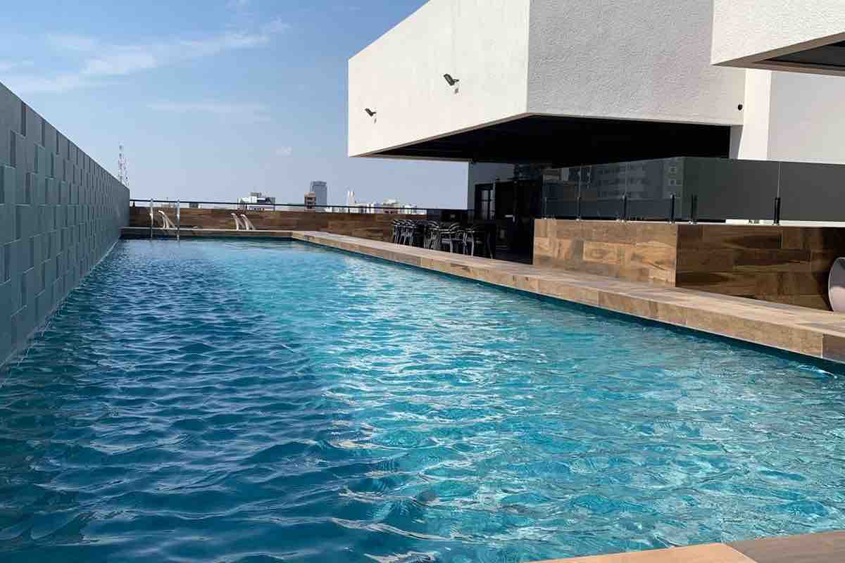 A clear blue pool stretches across a spacious deck, reflecting the sky above. Surrounding areas feature wooden accents, with seating visible in the shaded section nearby. The edge of the pool offers a seamless view of the city's skyline.
