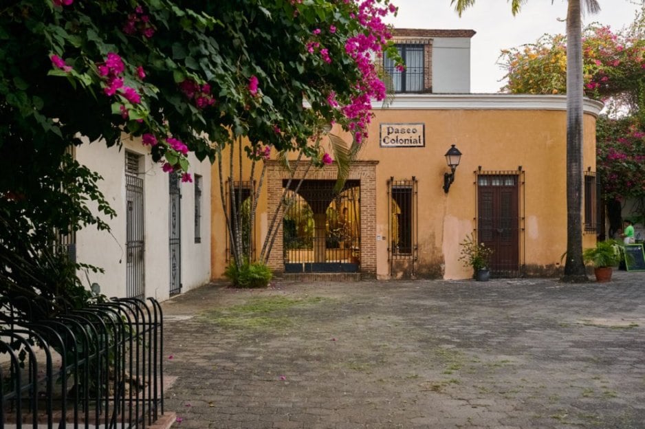 The entrance to the Paseo Colonial features a vivid display of flowering plants alongside a textured stone pathway. An arched passageway is framed by a rustic yellow wall. Decorative lighting adorns the facade, adding character to the inviting atmosphere.