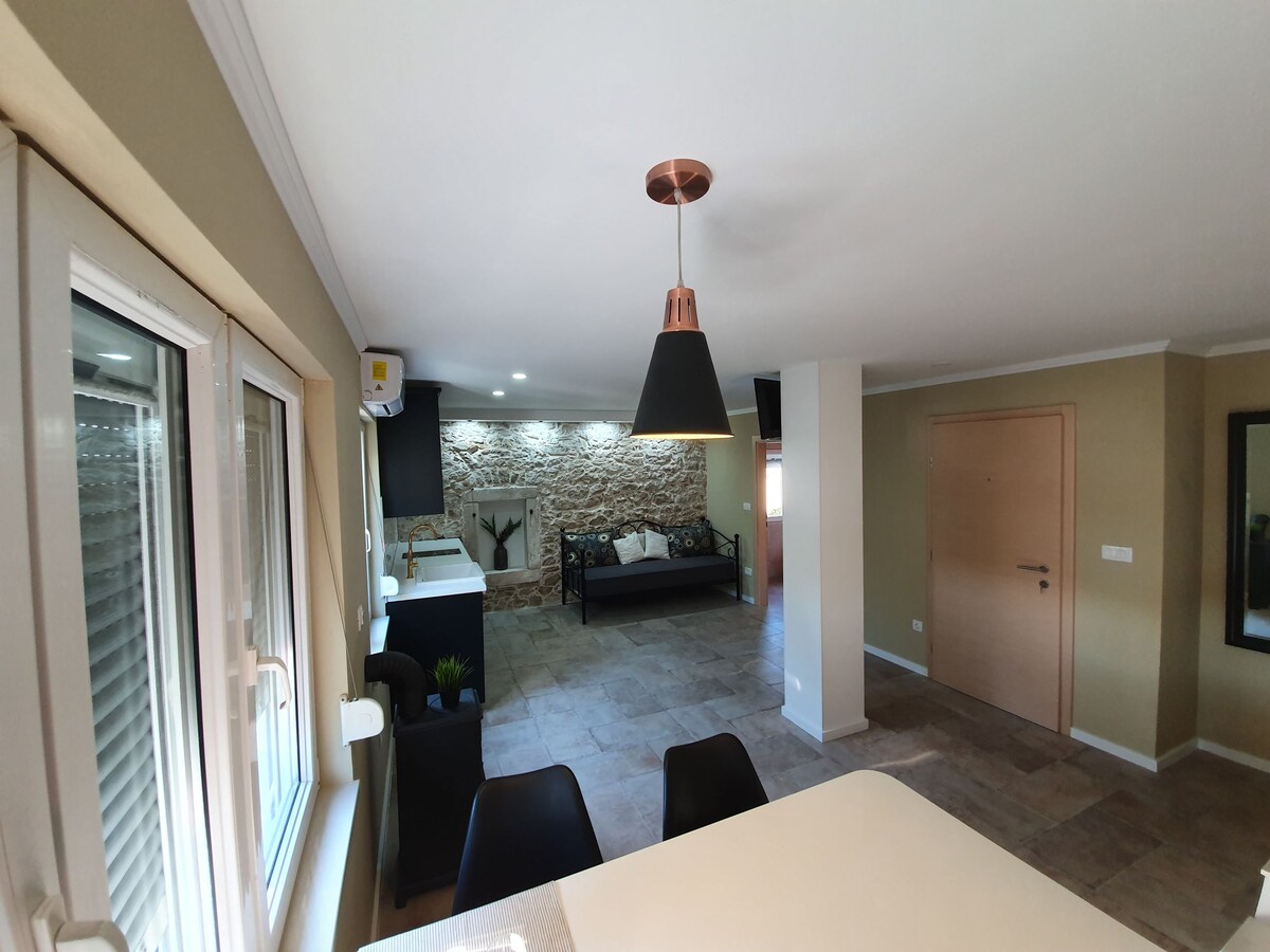 An open living space features a stylish kitchen area with modern fixtures. A cozy seating arrangement includes a dark colored sofa, while natural stone accent walls provide texture. Light-colored walls and large windows contribute to a bright and spacious atmosphere.