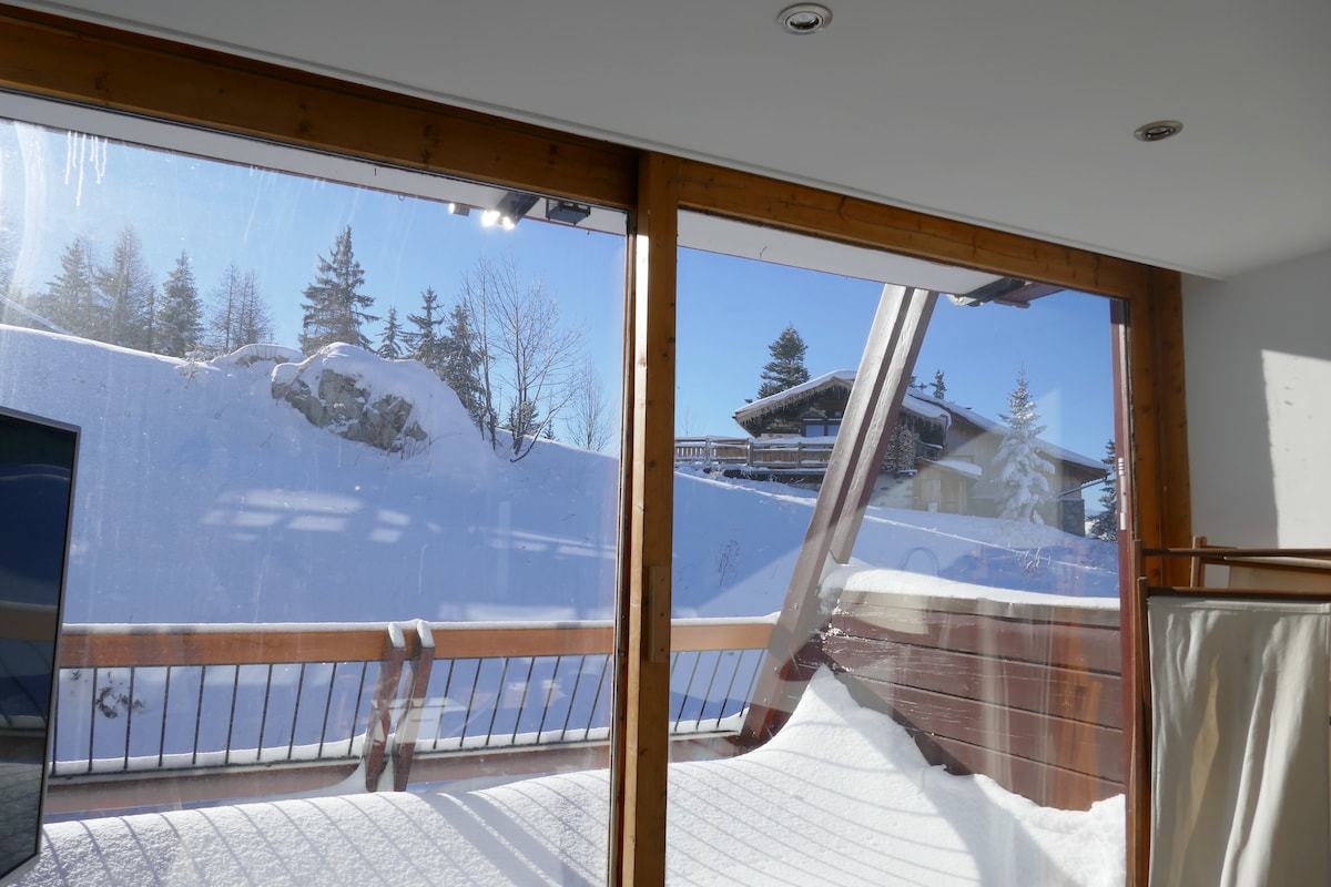 A view from the apartment's balcony is presented, showcasing a snowy landscape with trees and mountain lodges visible beyond. Sunlight streams in through large glass doors, enhancing the bright atmosphere of the space.