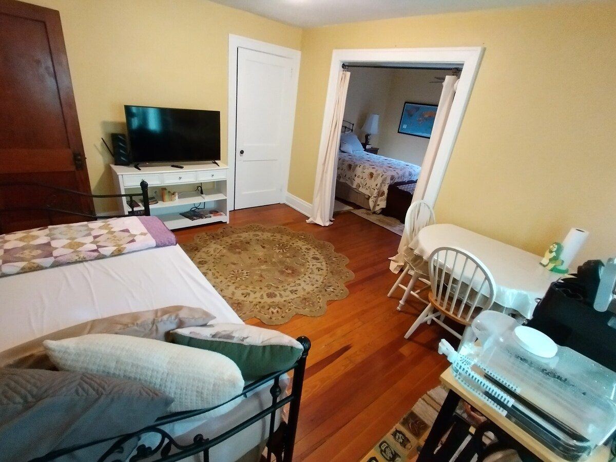 The room features a cozy layout with a black metal bed adorned in neutral tones. A small table with two chairs is positioned near a window. A mounted flat-screen TV is visible, along with a closed door leading to another bedroom. The warm yellow walls enhance the ambiance.