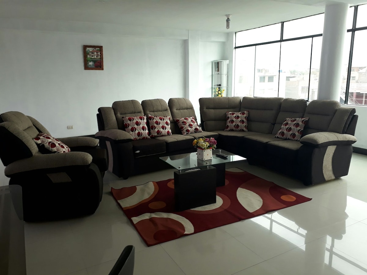 A modern living area is presented, featuring a spacious sectional sofa adorned with patterned cushions. A coffee table sits at its center, complemented by a vibrant area rug. Large windows allow ample natural light to illuminate the room.