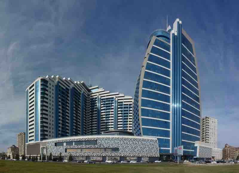 A modern high-rise building features a distinctive, curved glass facade, reflecting the sky in shades of blue. The structure is accompanied by a series of lower, multi-story sections with horizontal lines, presenting a contemporary architectural style. Surrounding landscaping includes grassy areas and pathways.
