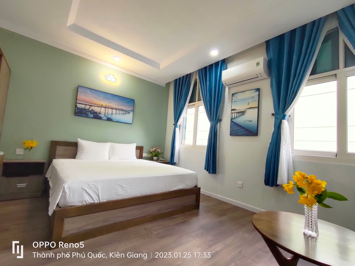 A bright and spacious room features a large bed with crisp white linens. Light walls are complemented by vibrant blue curtains framing two windows. A small bouquet of yellow flowers adds a touch of color to the wooden nightstand, while a serene piece of wall art hangs above the bed.
