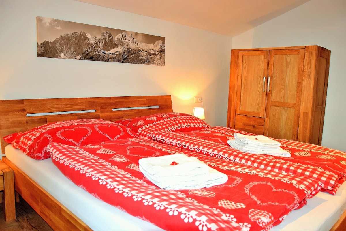 A cozy bedroom features a spacious bed adorned with red bedding, showcasing a heart pattern. Fresh towels are neatly arranged on the bed. A wooden wardrobe stands nearby, while a scenic mountain photograph is displayed above the headboard, contributing to a calm ambiance.