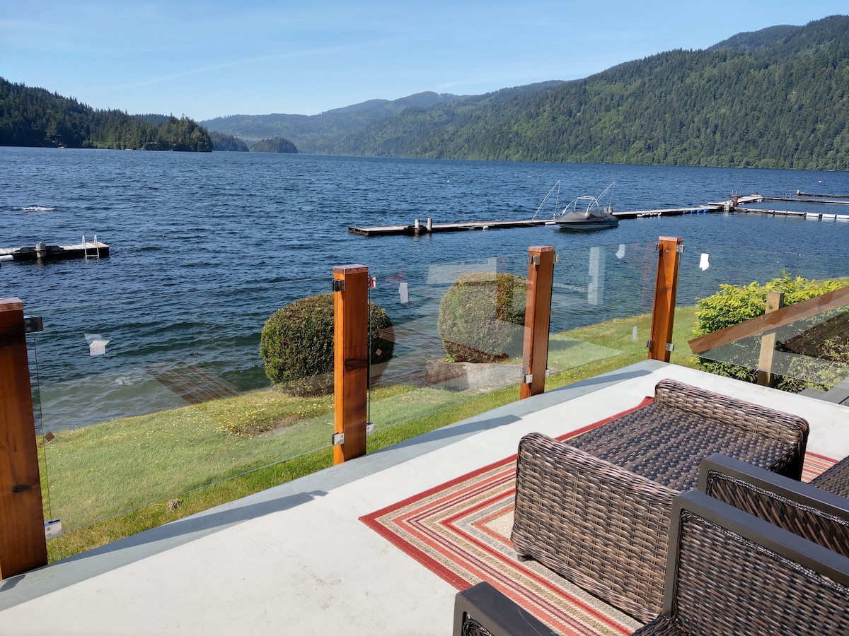 A tranquil outdoor seating area is positioned on a spacious patio, featuring woven seating and a patterned rug. Clear views of Lake Whatcom and surrounding mountains can be observed, with a grassy area leading to the water's edge and a wooden dock visible in the distance.