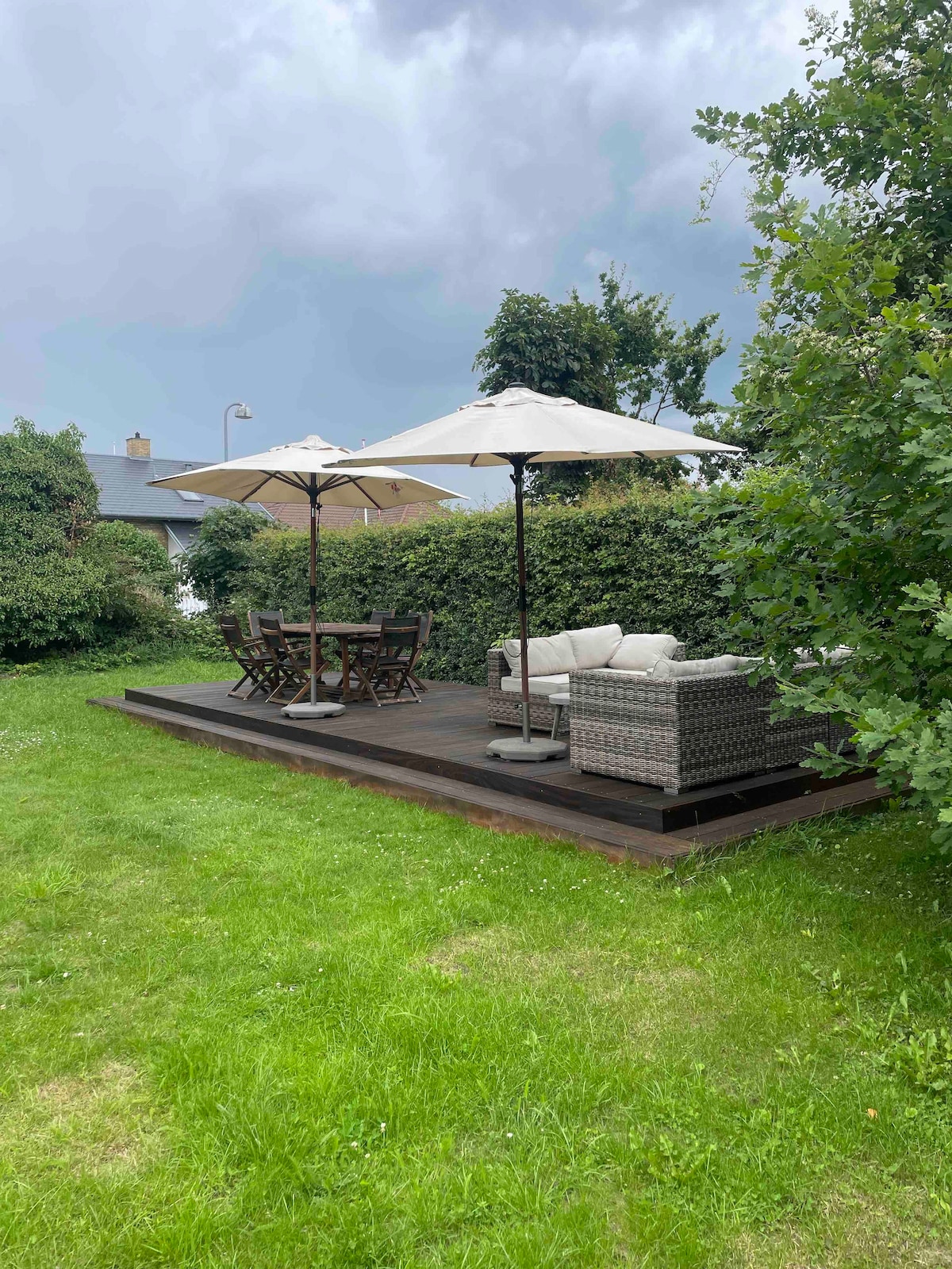 A wooden terrace is set within a lush garden, featuring lounge furniture under two umbrellas, providing shaded seating. A table with four chairs complements the seating area, surrounded by green grass and hedges, creating a comfortable outdoor space.