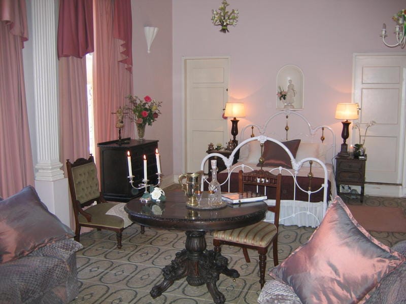 The family room features soft pink walls and elegant drapes, creating a soothing environment. A wrought-iron bed is positioned against the wall, complemented by a round dining table surrounded by upholstered seating. Warm light emanates from decorative lamps, enhancing the inviting decor.