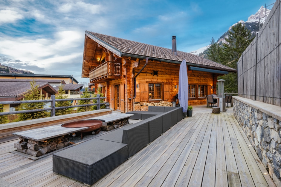 An outdoor deck area features a spacious seating arrangement made of dark, modern materials, with a circular fire pit at its center. The wooden chalet stands in the background, framed by the mountains and a clear sky, enhancing the inviting outdoor space.