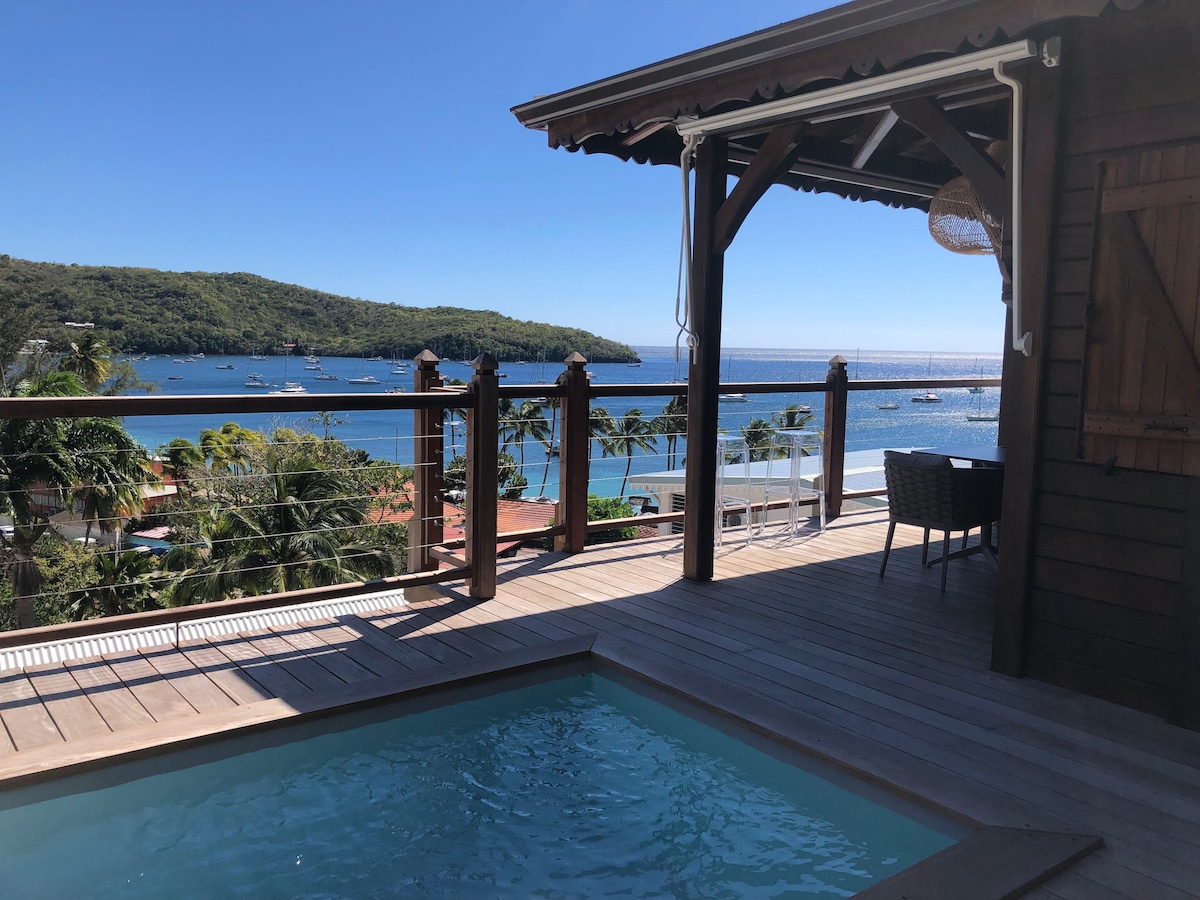 A private terrace with wooden decking features a small pool, offering views of the bay and surrounding hills. A table and chairs are positioned nearby, creating a serene outdoor space. Bright blue skies and calm waters enhance the relaxing atmosphere.