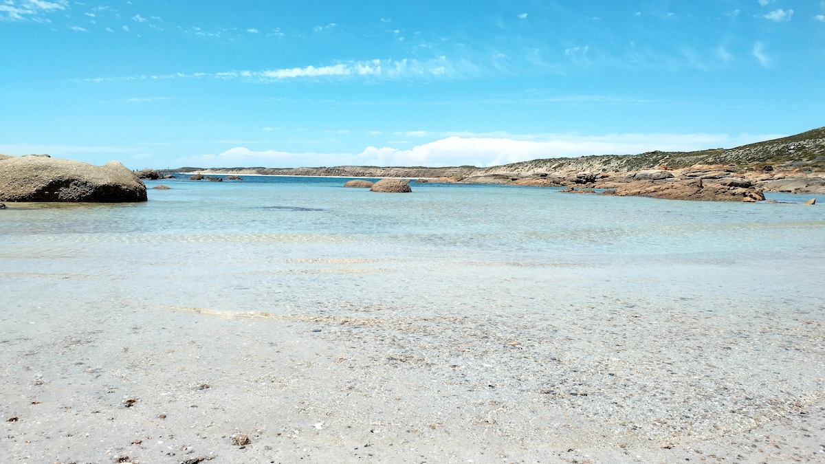 A serene shoreline presents gentle turquoise waters meeting a sandy beach. Smooth rocks are visible in the calm water, contributing to the tranquil seaside landscape under a clear blue sky with scattered clouds.