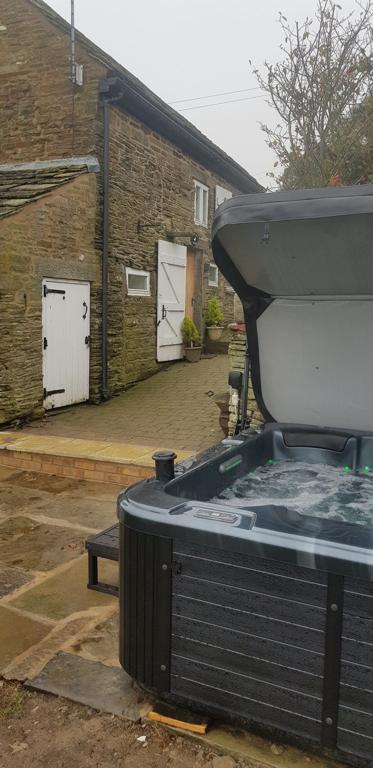 A hot tub is positioned on a paved area, showcasing jets of water and green lighting. The stone cottage provides a rustic backdrop, with a white door and window visible. A quiet, natural setting is evident, hinting at the tranquility of the surroundings.