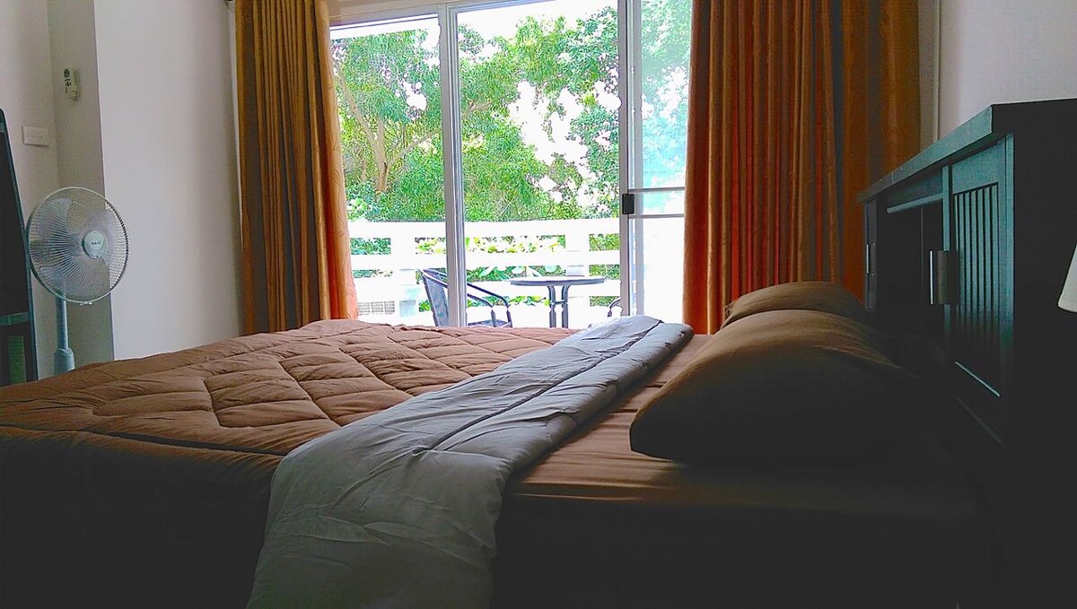 A spacious bedroom features a large bed with neutral-toned bedding. A small balcony is visible through the large glass doors, framed by light curtains. A fan is positioned near the bed, and the warm orange curtains enhance the natural light filtering into the room.
