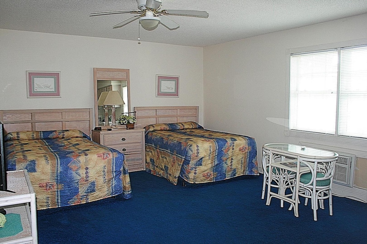 The spacious Studio Villa features two double beds with colorful bedding against a neutral wall. A large mirror is mounted above a wooden dresser. A small dining table with two chairs is positioned near a window that allows natural light to fill the room.