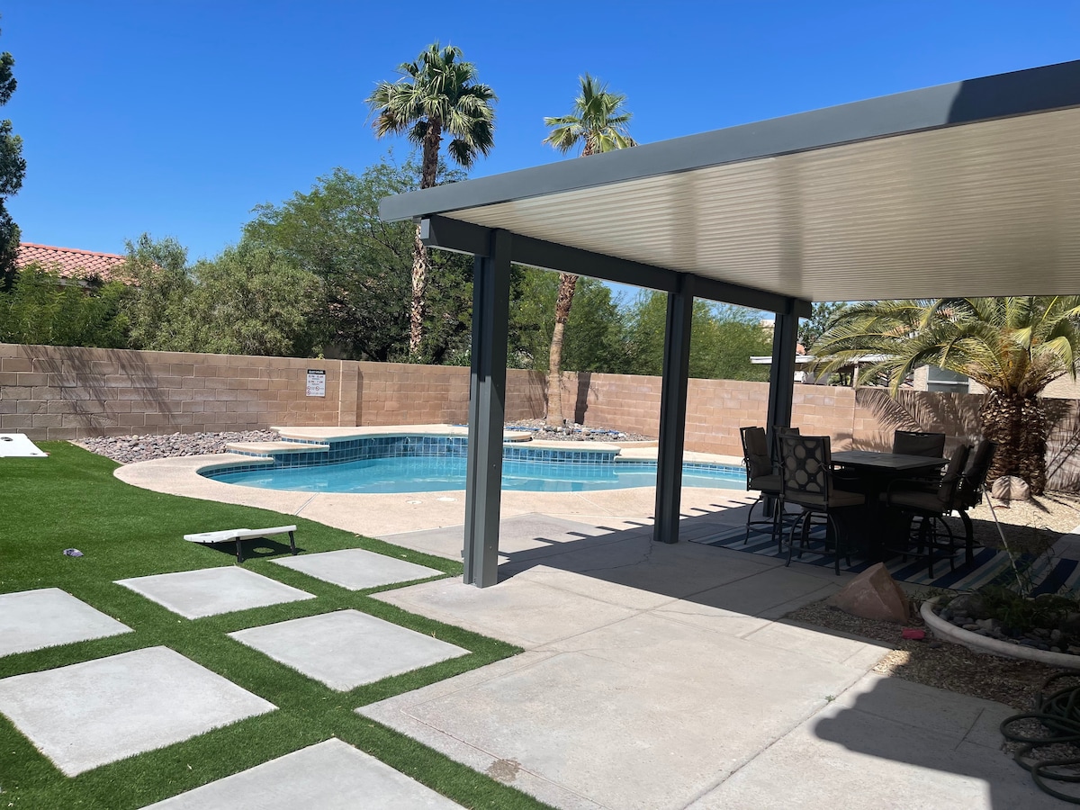 A spacious backyard is shown, featuring a private pool surrounded by a sun patio. A covered seating area with a dining table is positioned nearby, and tropical plants add greenery to the landscape. Pathways of square pavers are visible leading to the pool area.