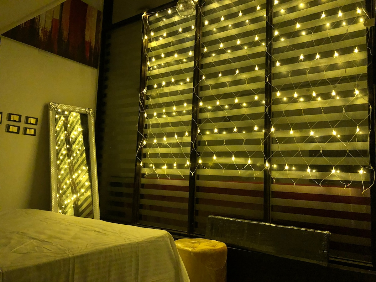 A softly lit interior showcases a spacious bed covered with a white linen. The backdrop features glowing string lights cascading down sheer curtains, creating a warm ambiance. A large mirror reflects the light, while a small, round ottoman adds a pop of color to the room.