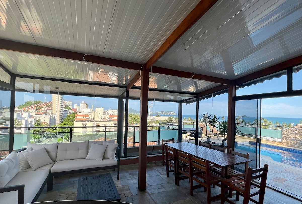 An inviting covered terrace showcases a spacious dining area with wooden tables and chairs. Large glass windows offer panoramic views of the surrounding landscape and ocean. A comfortable seating arrangement is positioned nearby, and a private pool is visible in the background, enhancing the relaxed ambiance.