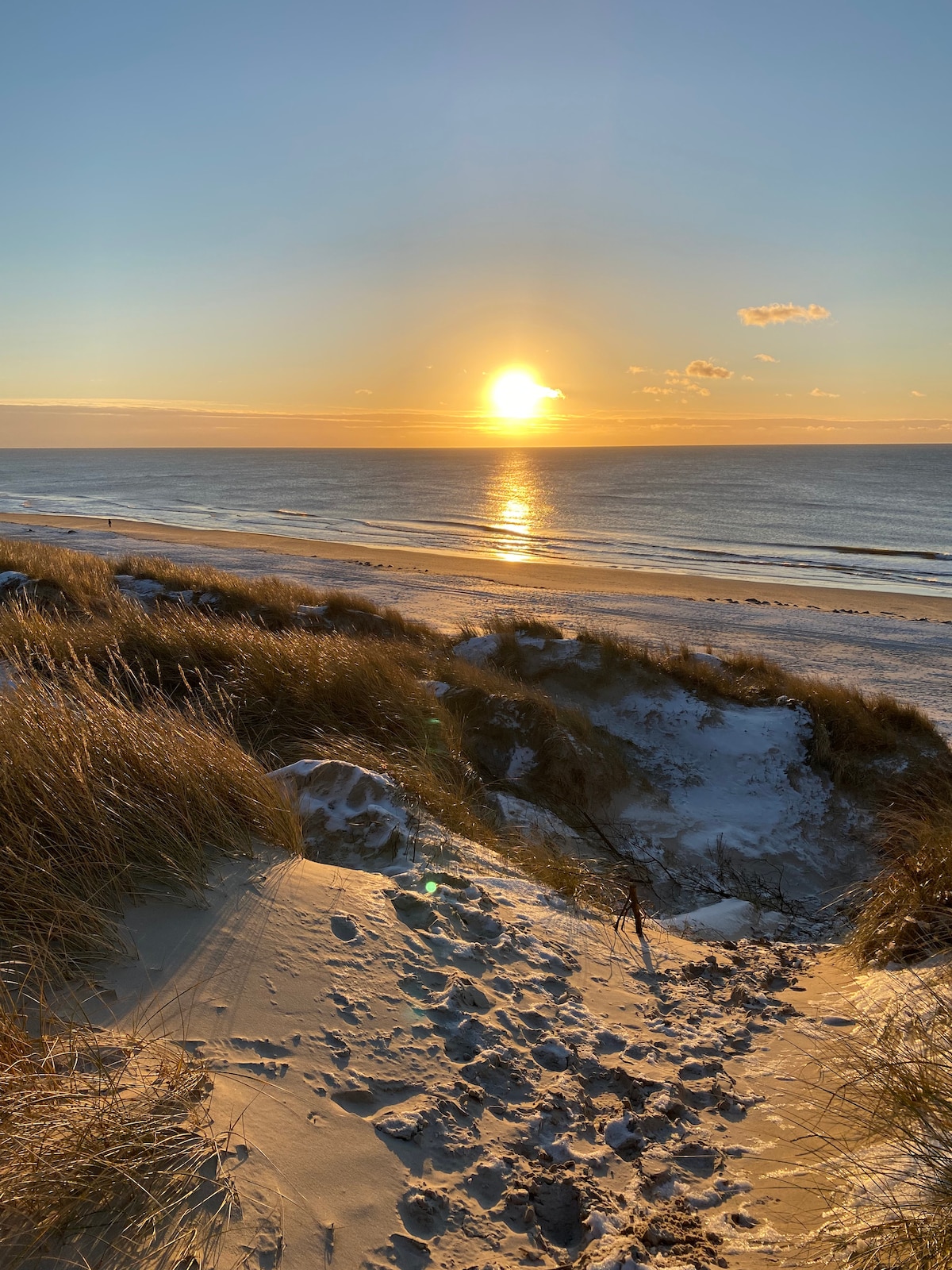 A serene beach scene captures the sun setting over calm waters, creating a reflective glow on the surface. Golden sand dunes covered with grass lead down to the shore, where the beach is visible. The sky is painted with soft hues of orange and blue.
