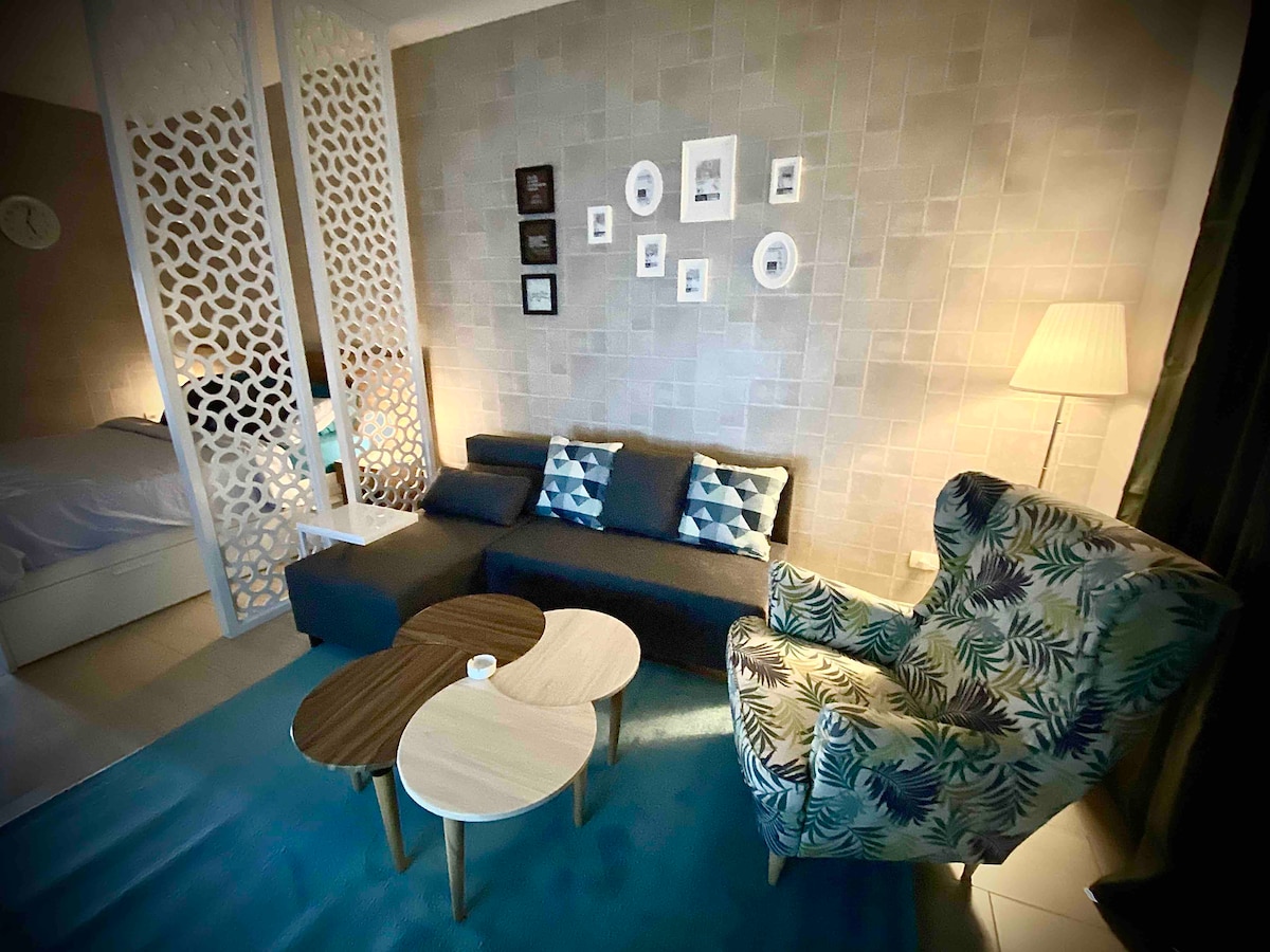 A cozy living area is highlighted by a stylish sofa adorned with blue and white cushions. A patterned armchair complements the space, while a pair of wooden coffee tables sit center stage on a soft rug. Subtle wall decor adds a touch of personalization.